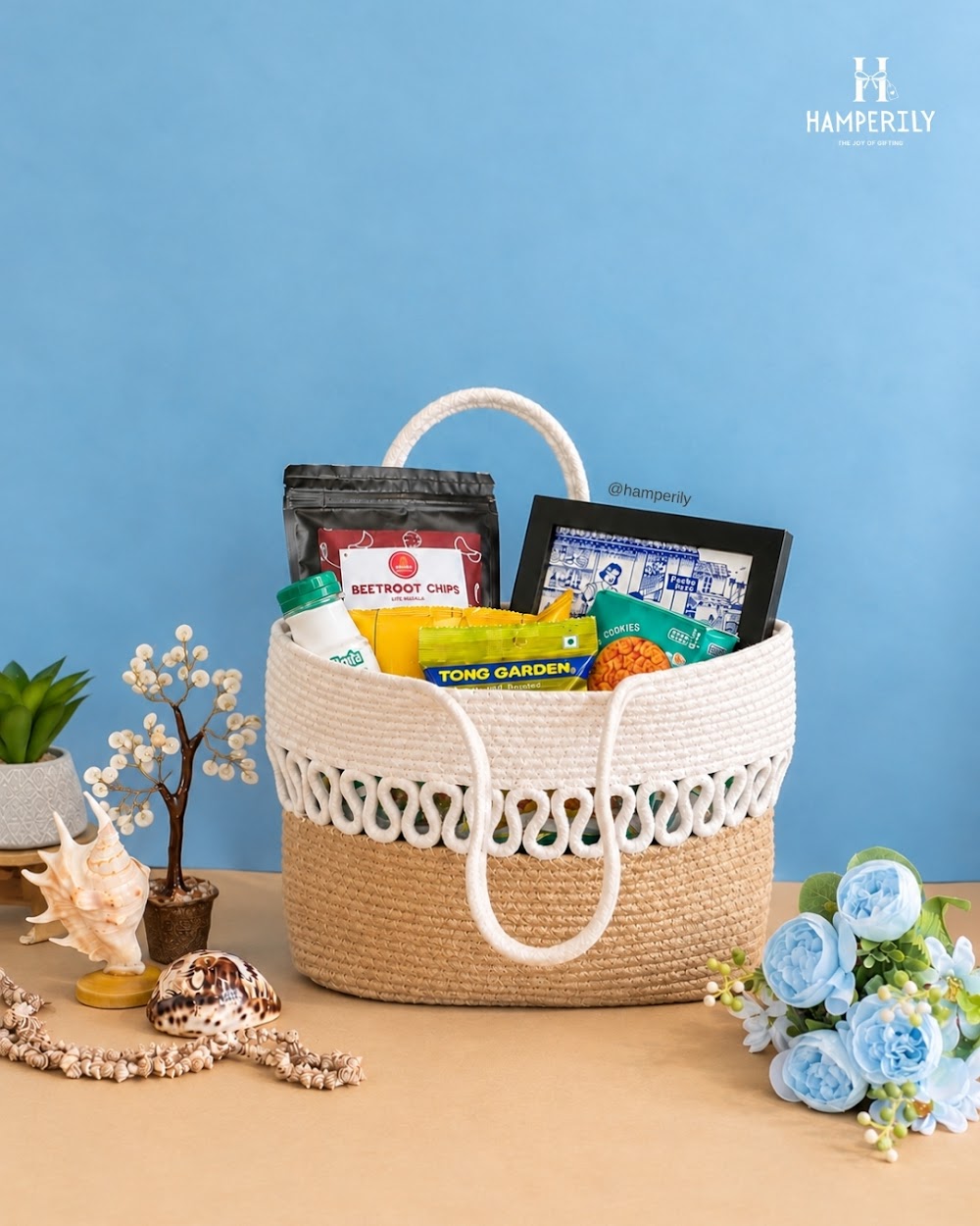 Hamperily rope basket hamper with snacks, flowers and shells on a blue backdrop